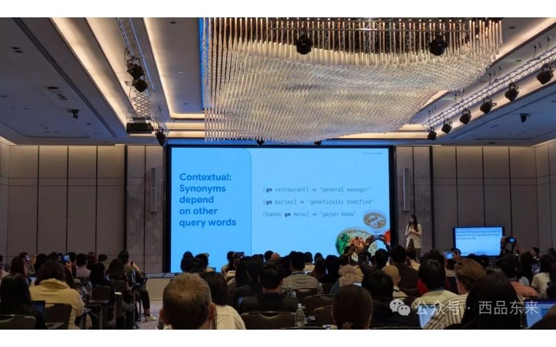 一个人正在会议室中做演讲，背景是投影屏幕，上面显示着英语文本，听众坐在下方。
