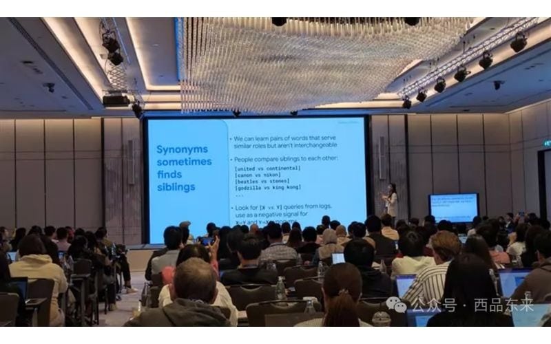 会议室内，一位讲师正在大屏幕前进行演讲，台下观众正在认真听讲和记录。